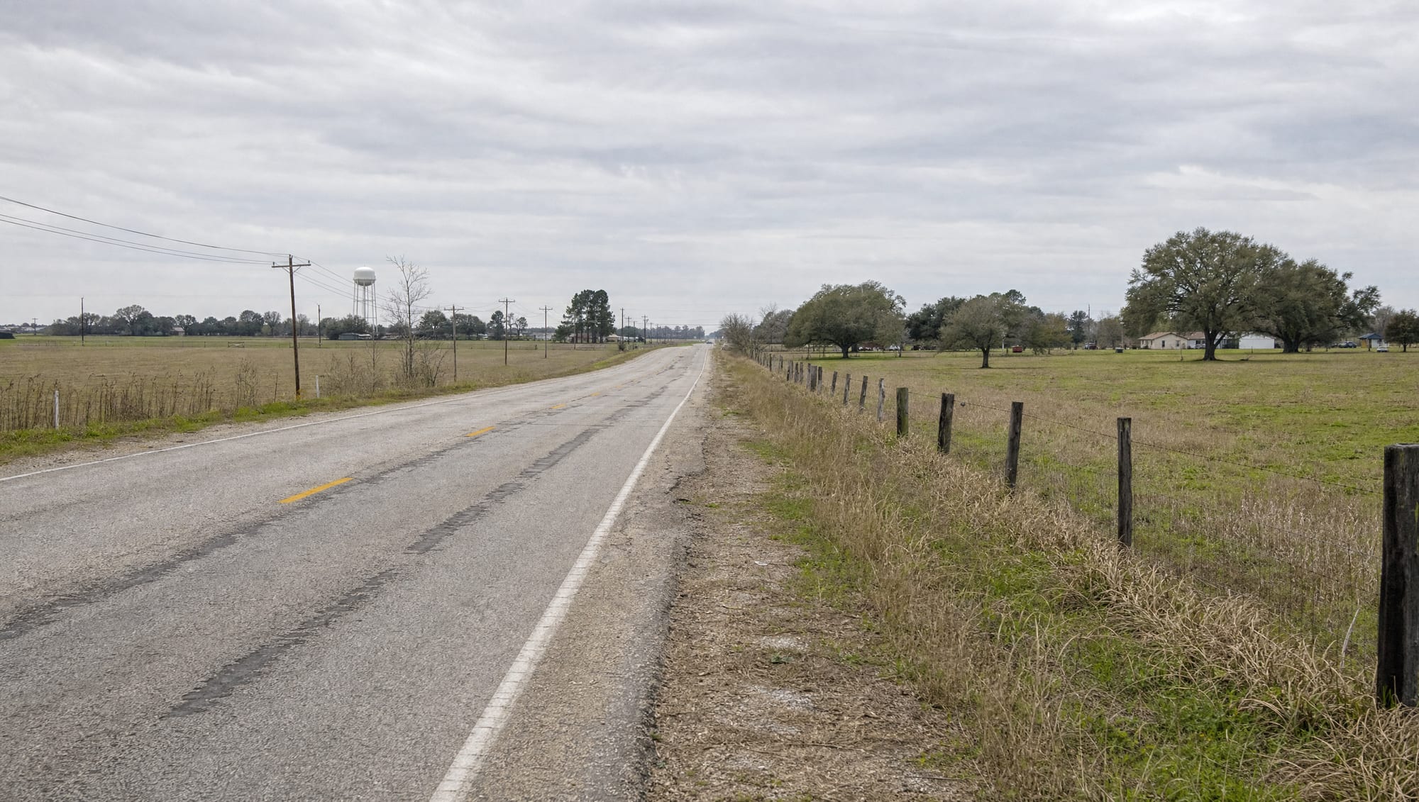 A quiet two-lane farm-to-market road running straight through flat open Fort Bend County farmland near Arcola, Texas, with a distant water tower, a wire-and-post fence line, scattered live oaks, and modest homesteads under a soft overcast sky