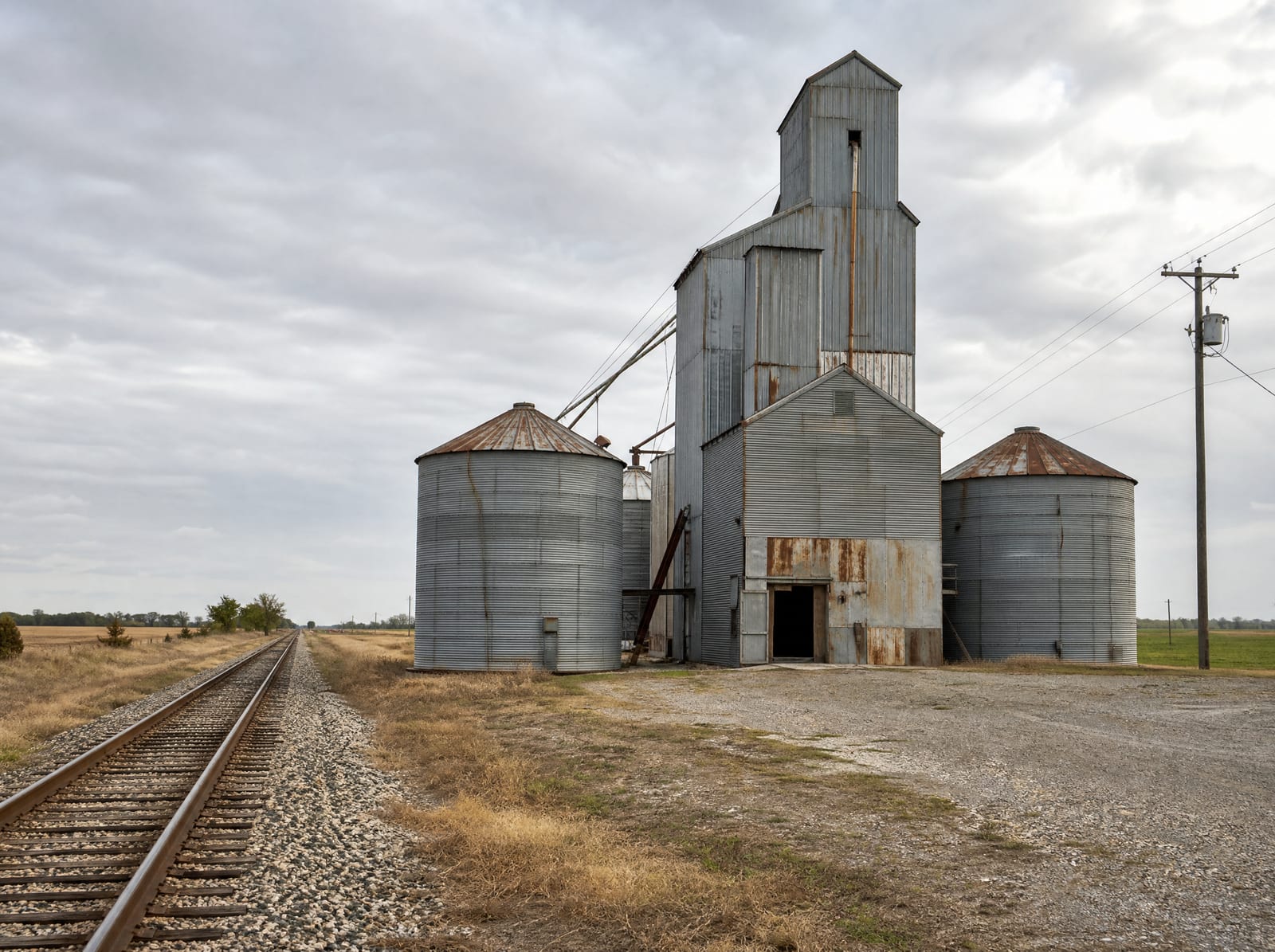 A weathered metal grain elevator and storage silos beside a single railroad track running through flat Fort Bend County farmland near Beasley, Texas, with a gravel lot and utility poles