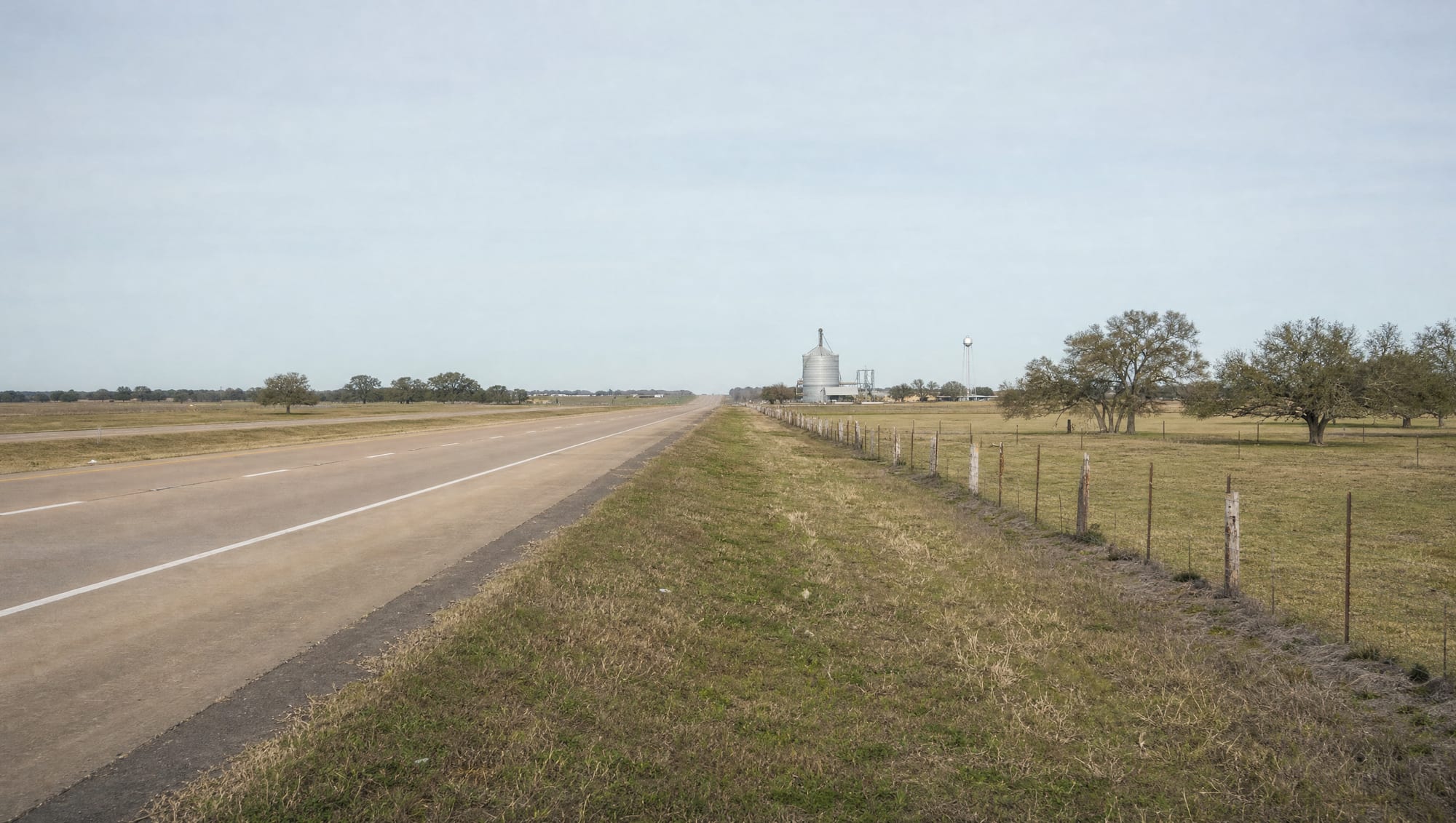 A wide highway corridor running through flat open Fort Bend County farmland and pasture near Beasley, Texas, with a distant grain silo and water tower, a wire fence, and scattered live oaks under a big pale sky