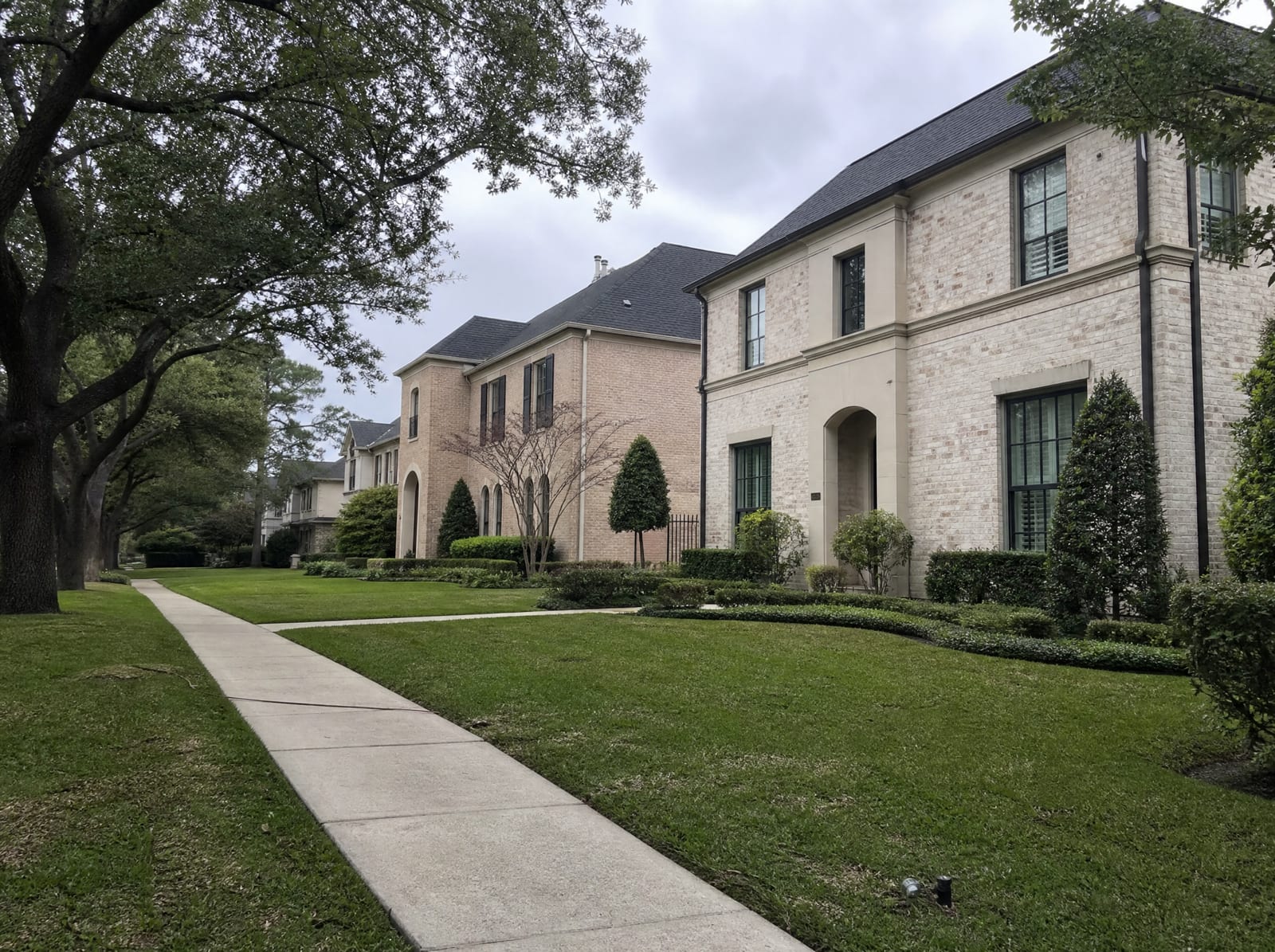 A quiet upscale residential street in Bellaire, Texas, with newer large two-story custom brick homes on modest urban lots, mature shade trees, manicured landscaping, and wide sidewalks under an overcast sky