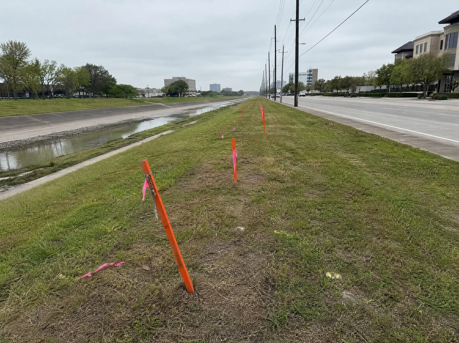 Orange survey stakes and pink right-of-way flags along a grassy strip beside a wide urban drainage channel and arterial in Bellaire, Texas, with utility poles, a concrete-lined bayou, and distant low-rise buildings, showing right-of-way acquisition pressure