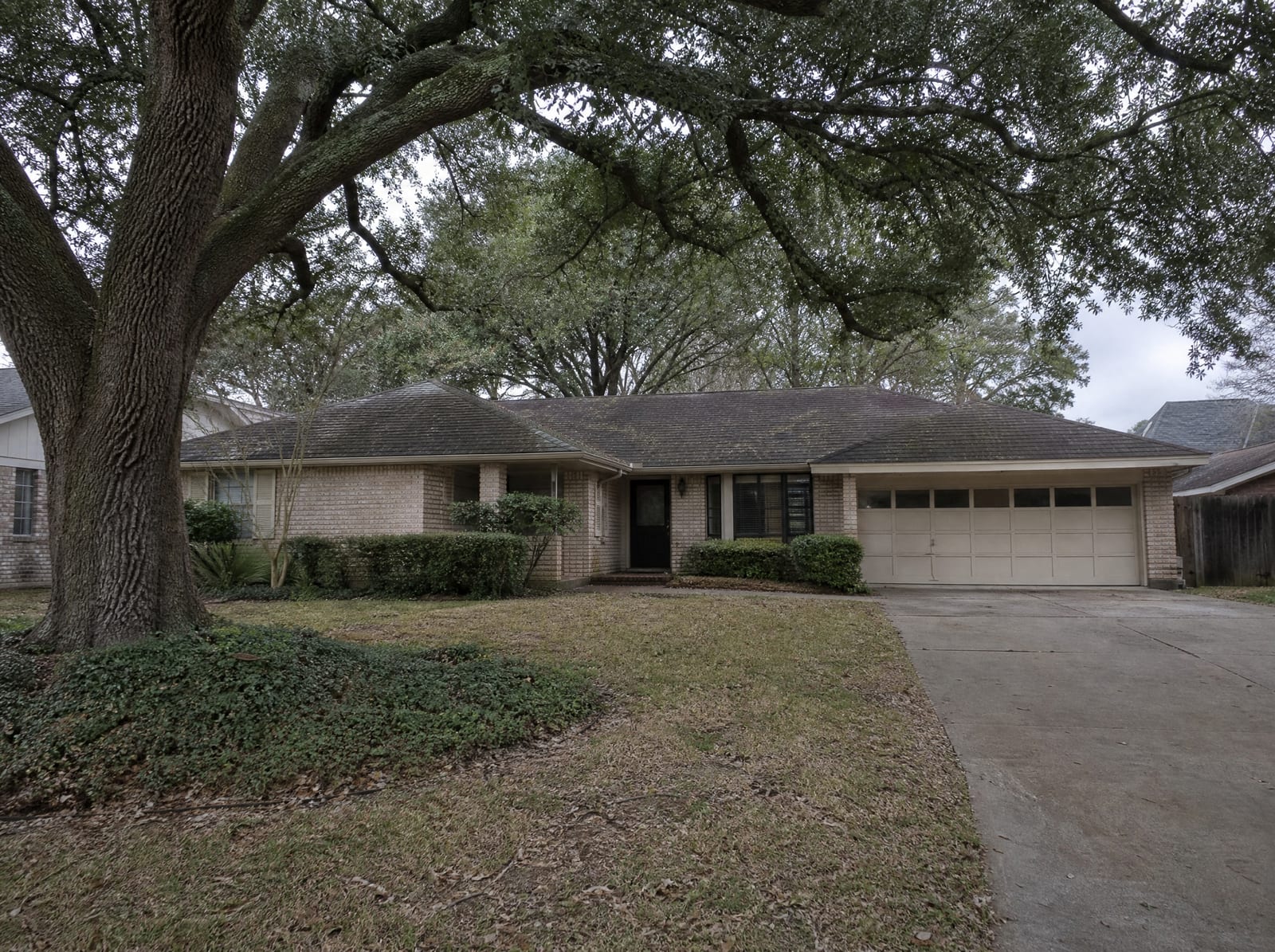 A classic older single-story mid-century ranch home on a shaded lot in an established Bellaire, Texas neighborhood, with a large mature live oak, a modest brick facade, and a front lawn under an overcast sky