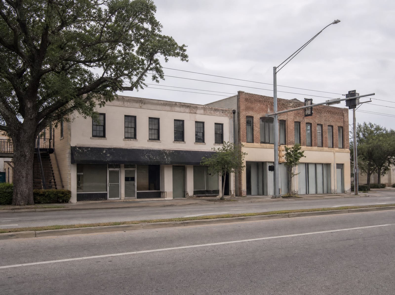 A low-rise urban commercial frontage along a busy boulevard in Bellaire, Texas near the Houston Loop, with modest mixed retail and office buildings, street trees, sidewalks, and traffic signals under an overcast sky