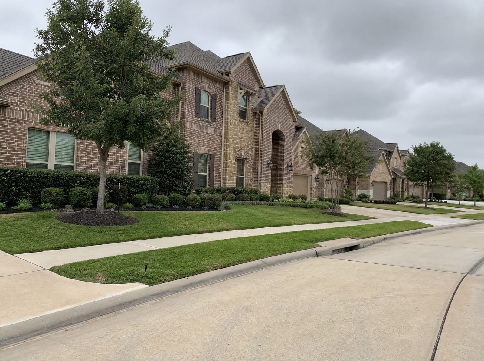 A quiet upscale master-planned residential street in Cinco Ranch near Katy, Texas, with larger two-story brick and stone homes, manicured landscaping, young trees, wide sidewalks, and a curbed street under an overcast sky
