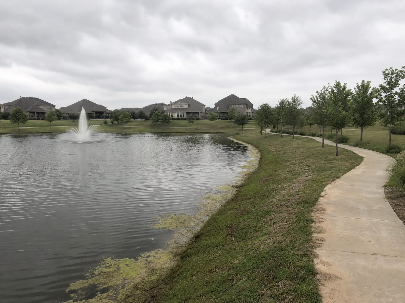 A landscaped community lake in Cinco Ranch near Katy, Texas, with manicured grassy banks, a paved walking trail, a small fountain, young trees, and master-planned suburban rooftops beyond under an overcast sky