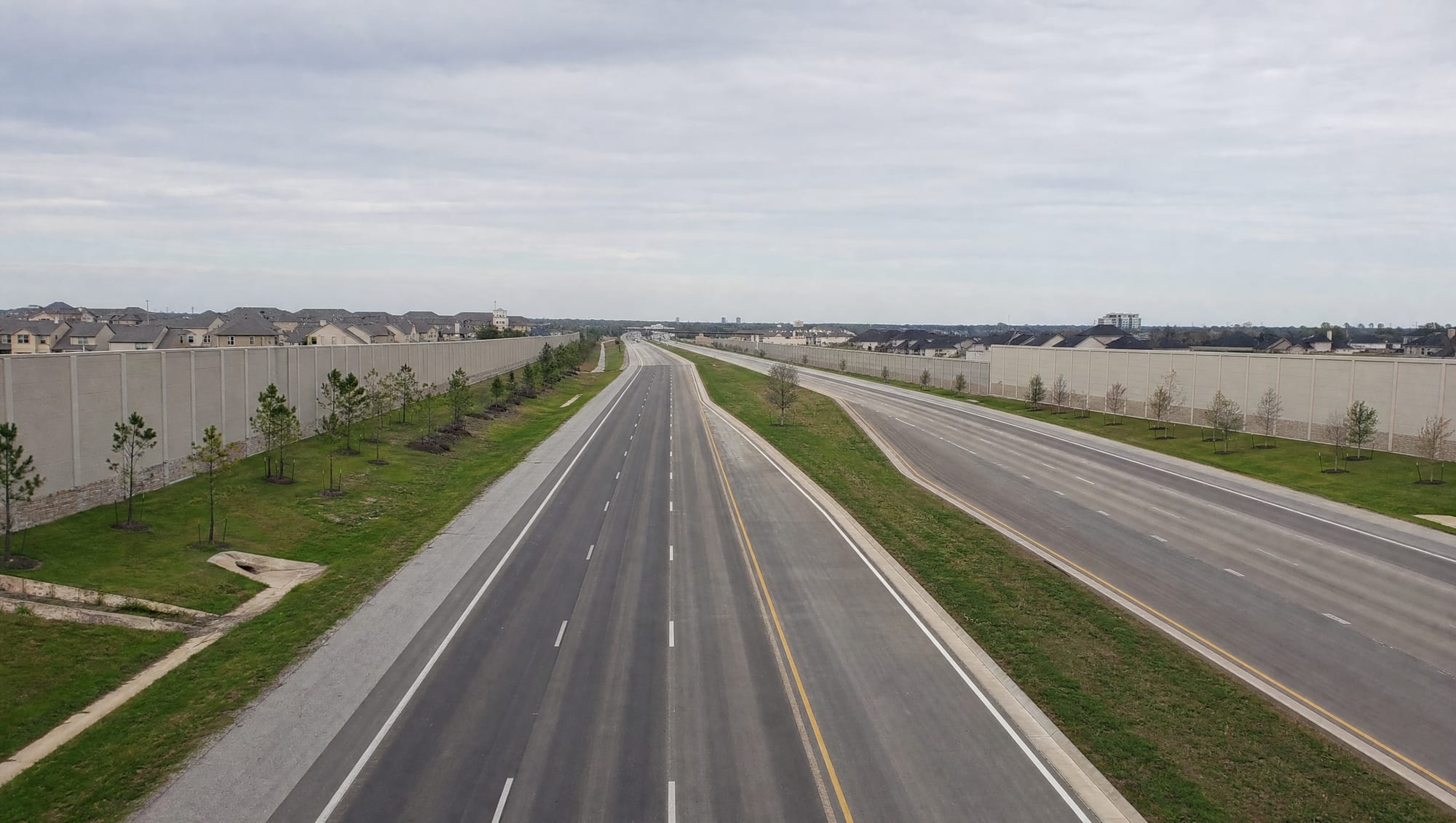 The Grand Parkway corridor near Cinco Ranch in the Katy, Texas area, a wide modern multi-lane highway with tall concrete sound walls, landscaped medians and frontage, young trees, and master-planned suburban development under a big pale sky