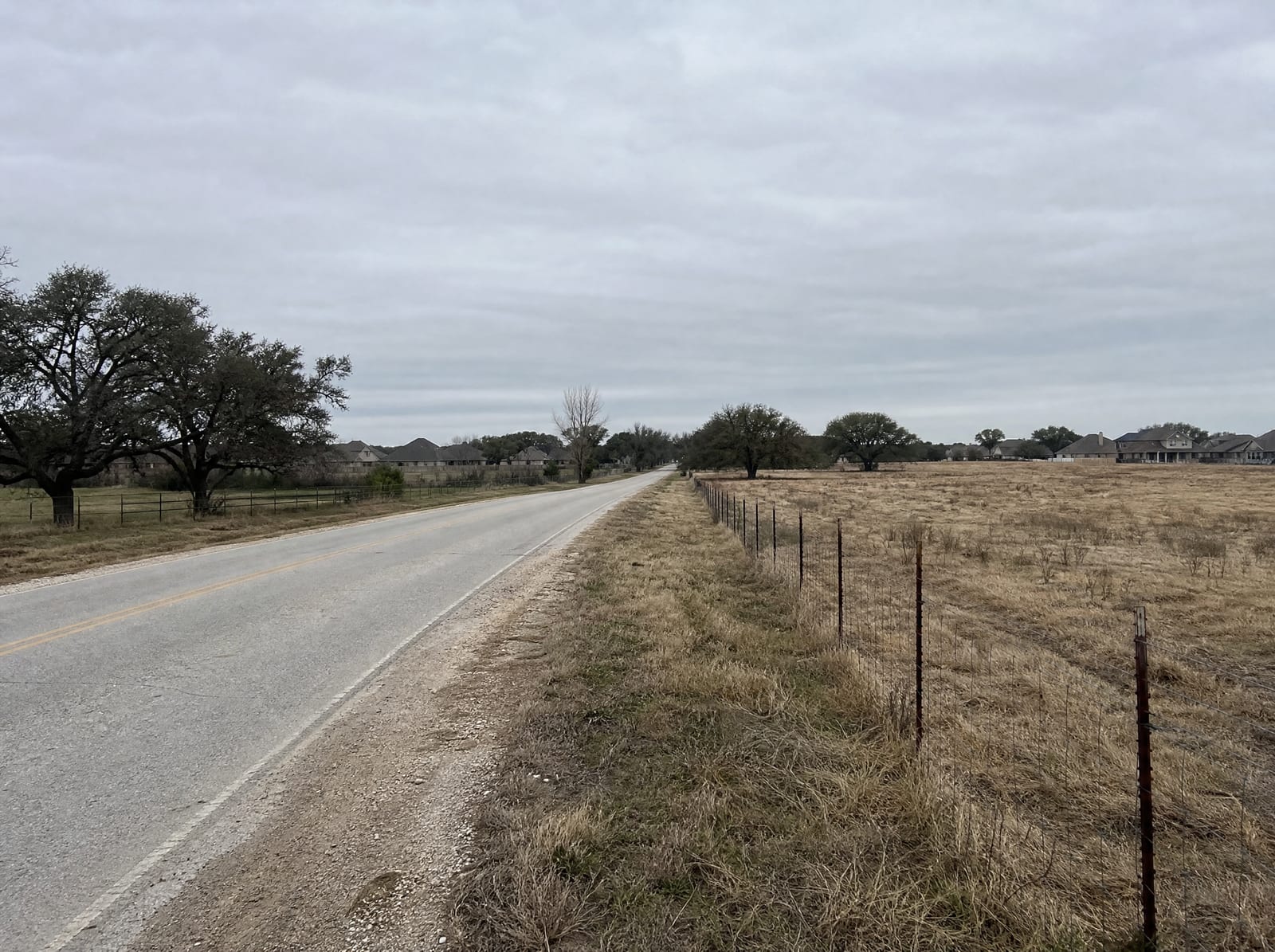 A quiet country road on the rural-to-suburban edge of Fulshear, Texas, with open pasture and a wire fence on one side and newer suburban rooftops emerging beyond scattered live oaks on the other, under a big pale overcast sky
