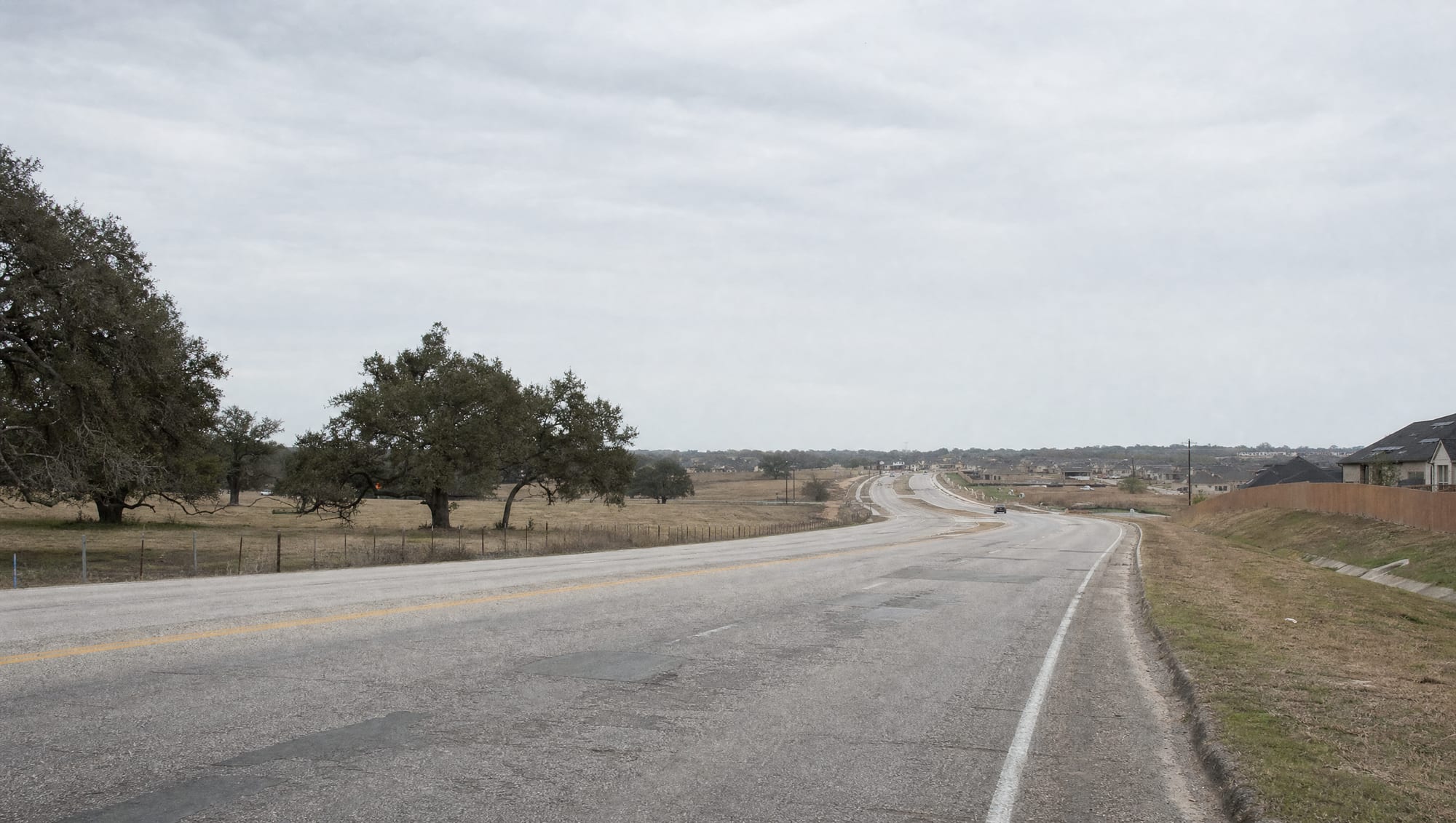 The FM 1093 and Westpark corridor near Fulshear, Texas in Fort Bend County, a widening road transitioning from open ranch land and pasture into newer master-planned suburban development, with scattered live oaks, a wire fence, and a big pale sky