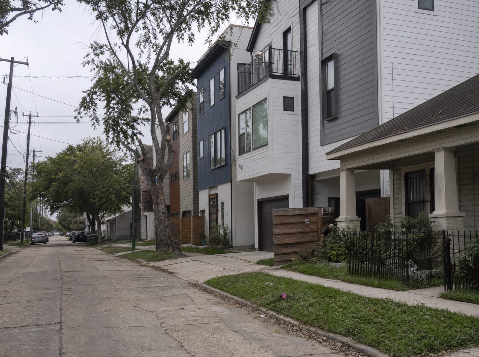 A dense inner-city Houston, Texas residential street with a mix of older one-story bungalows and newer three-story townhomes on narrow urban lots, mature shade trees, sidewalks, and utility poles under an overcast sky