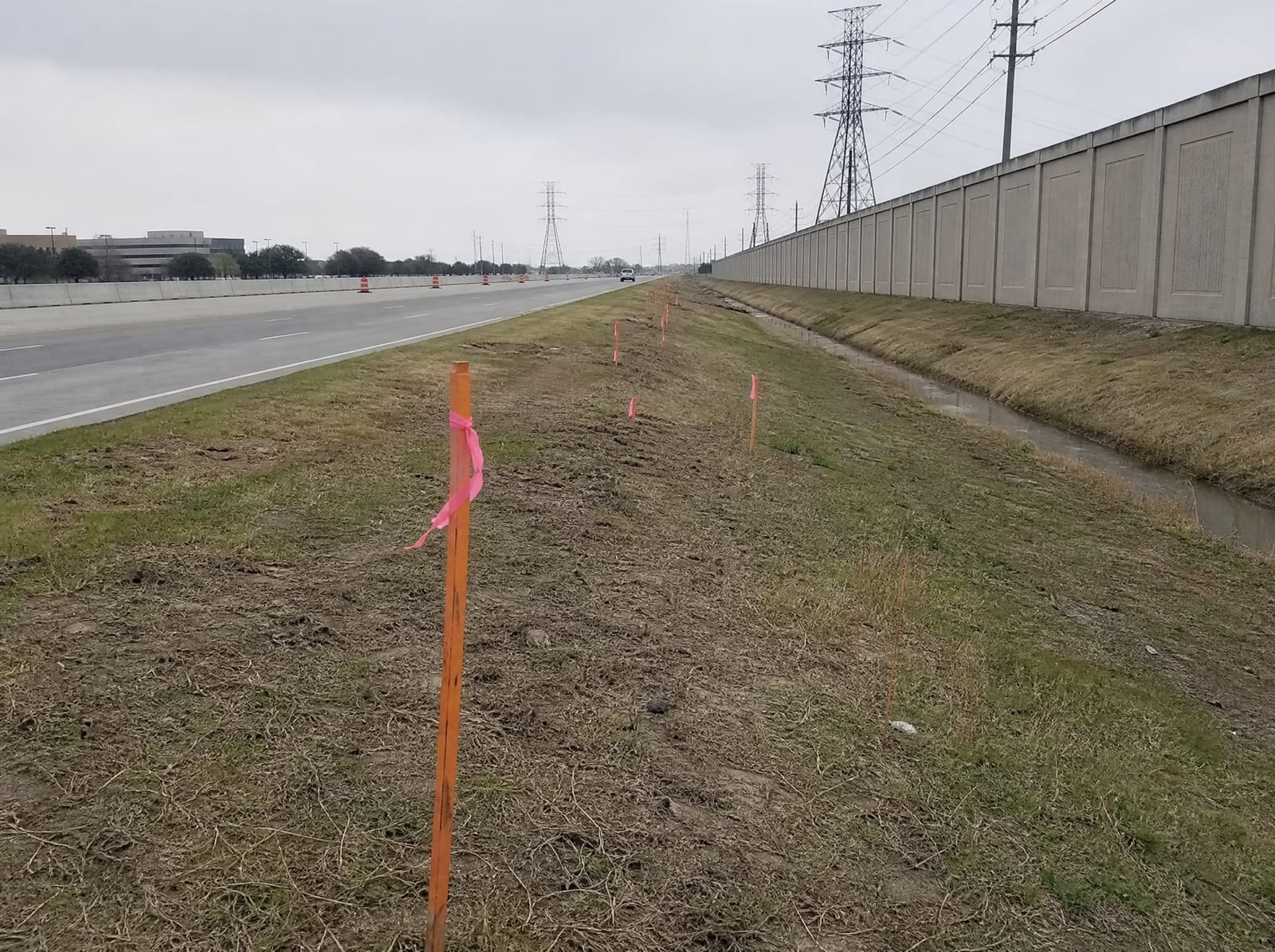 Orange survey stakes and pink right-of-way flags along a grassy strip beside a widening multi-lane urban freeway in Houston, Texas, with a tall concrete sound wall and distant transmission towers, showing right-of-way acquisition pressure