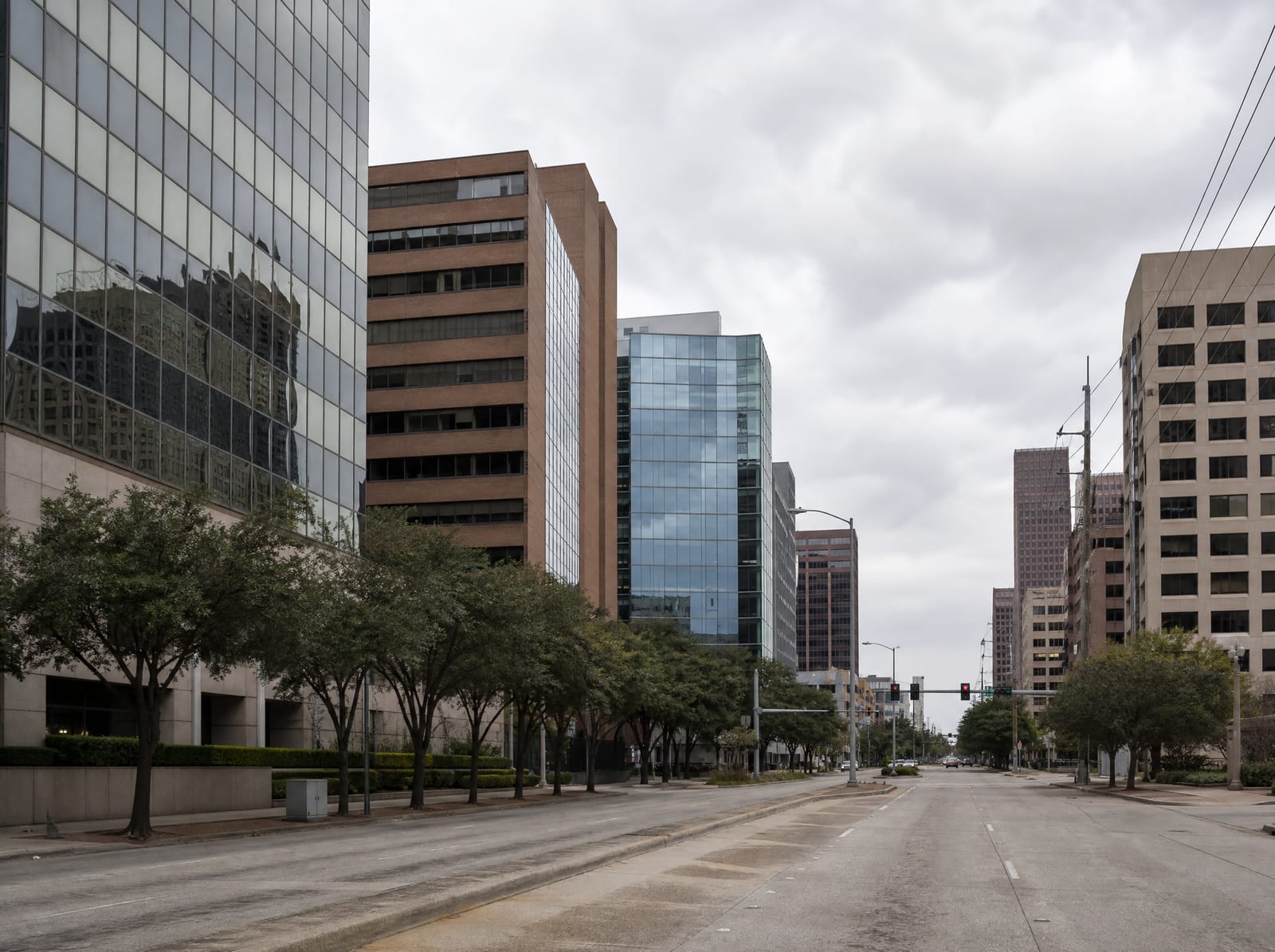 A mid-rise commercial district in Houston, Texas, with glass and brick mid-rise office buildings along a wide urban boulevard, street trees, sidewalks, and traffic signals under an overcast sky
