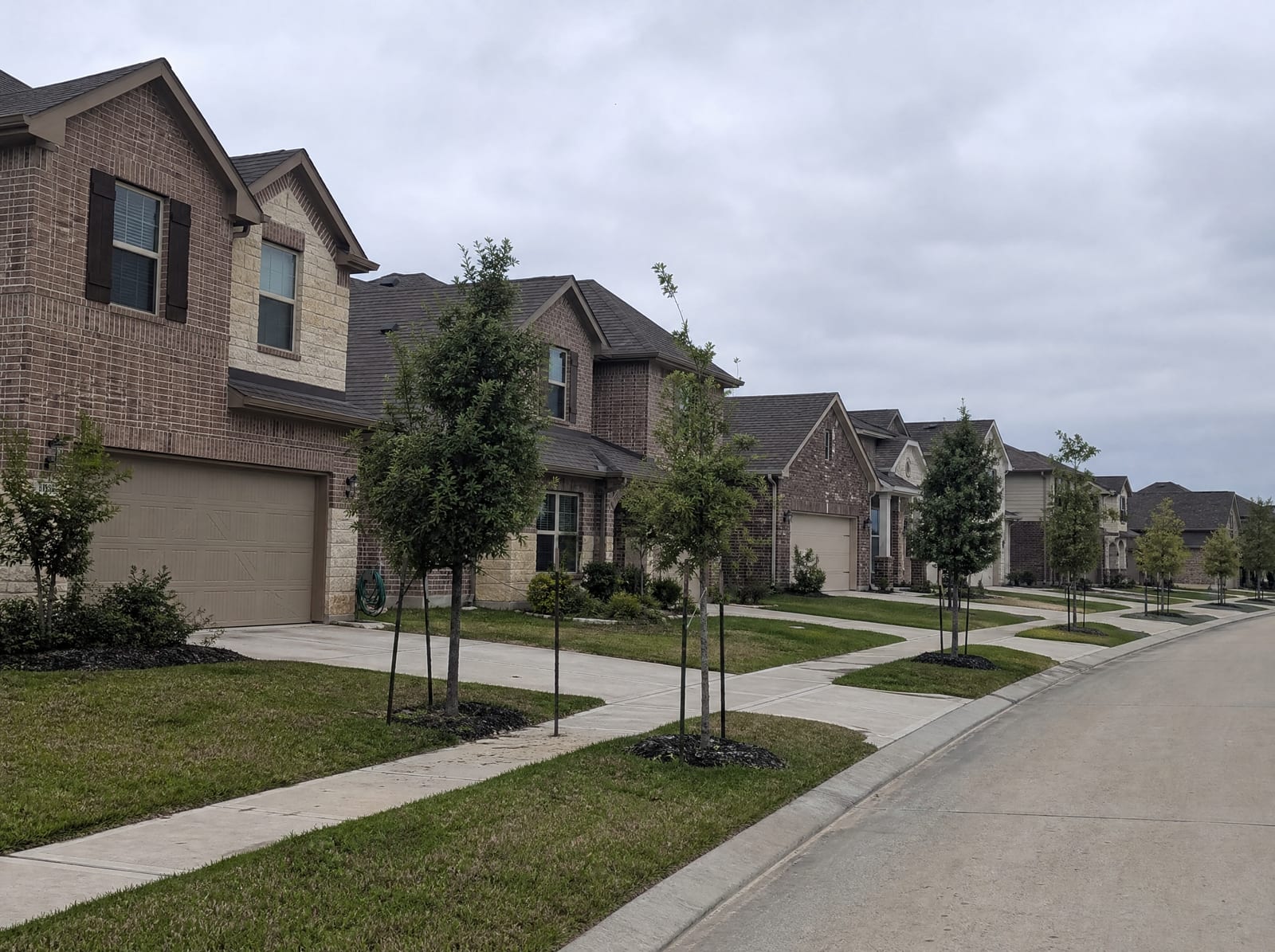 A typical Harris County suburban subdivision on the edge of Houston, Texas, with rows of two-story brick homes, manicured lawns, young landscaped trees, and wide sidewalks under an overcast sky