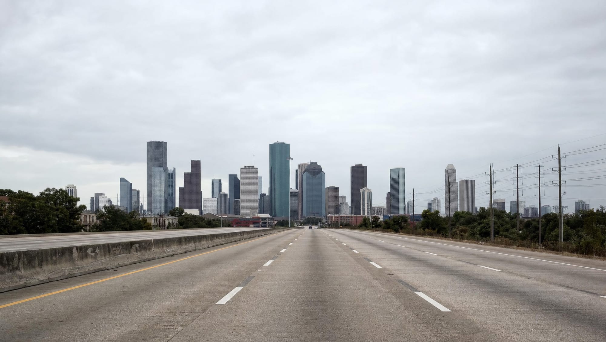 The downtown Houston, Texas skyline of glass and steel high-rise towers seen across a wide multi-lane urban freeway with a concrete interchange and overpasses in the foreground under a big pale overcast sky