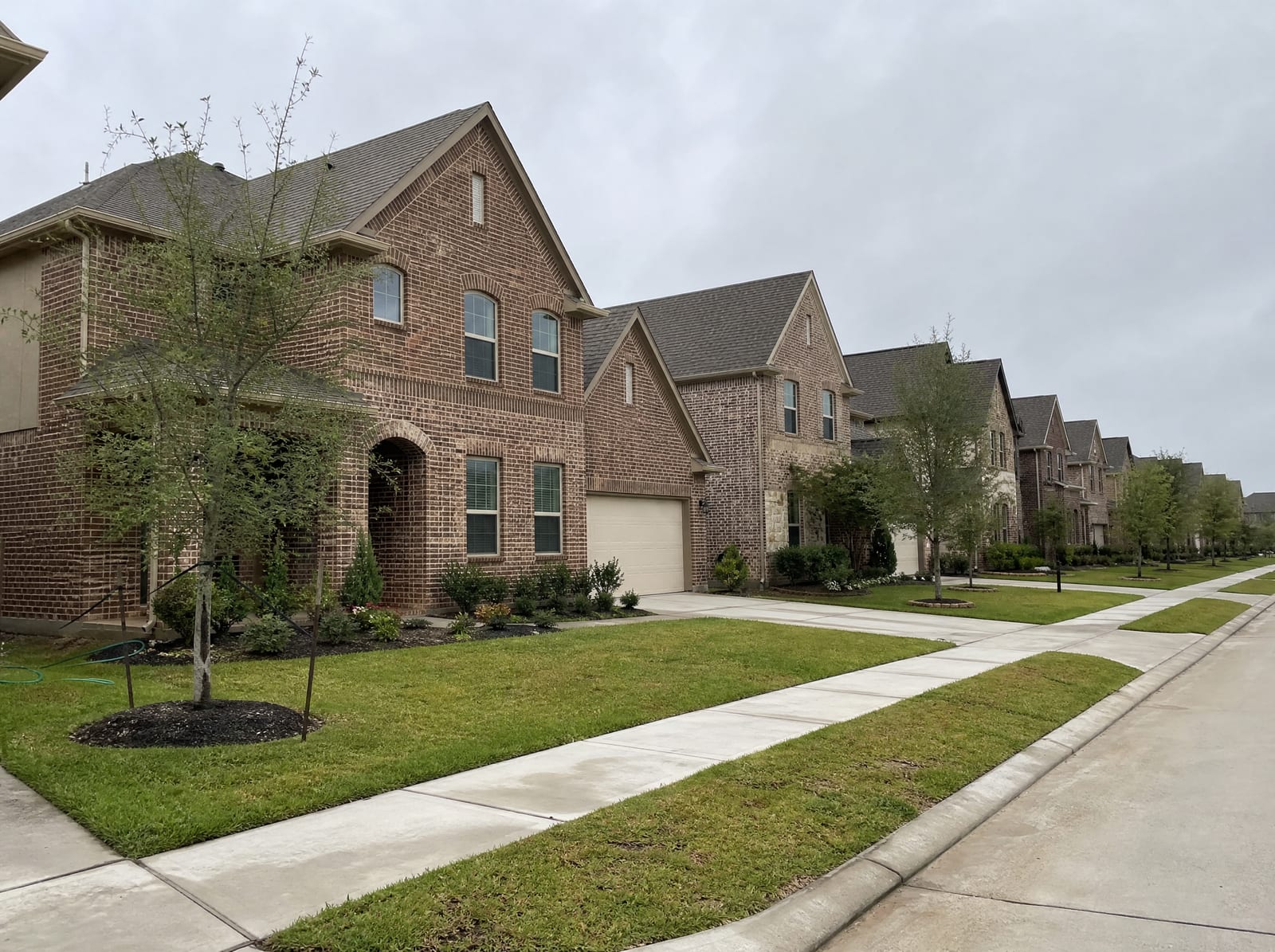 A quiet master-planned residential street in Katy, Texas, with two-story brick suburban homes, manicured lawns, young landscaped trees, wide sidewalks, and a curbed street under an overcast sky