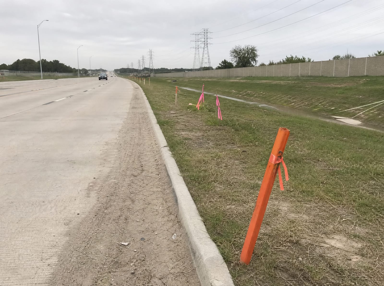 Orange survey stakes and pink right-of-way flags along a grassy strip beside a widening multi-lane suburban arterial near Katy, Texas, with a concrete sound wall and distant transmission towers, showing right-of-way acquisition pressure