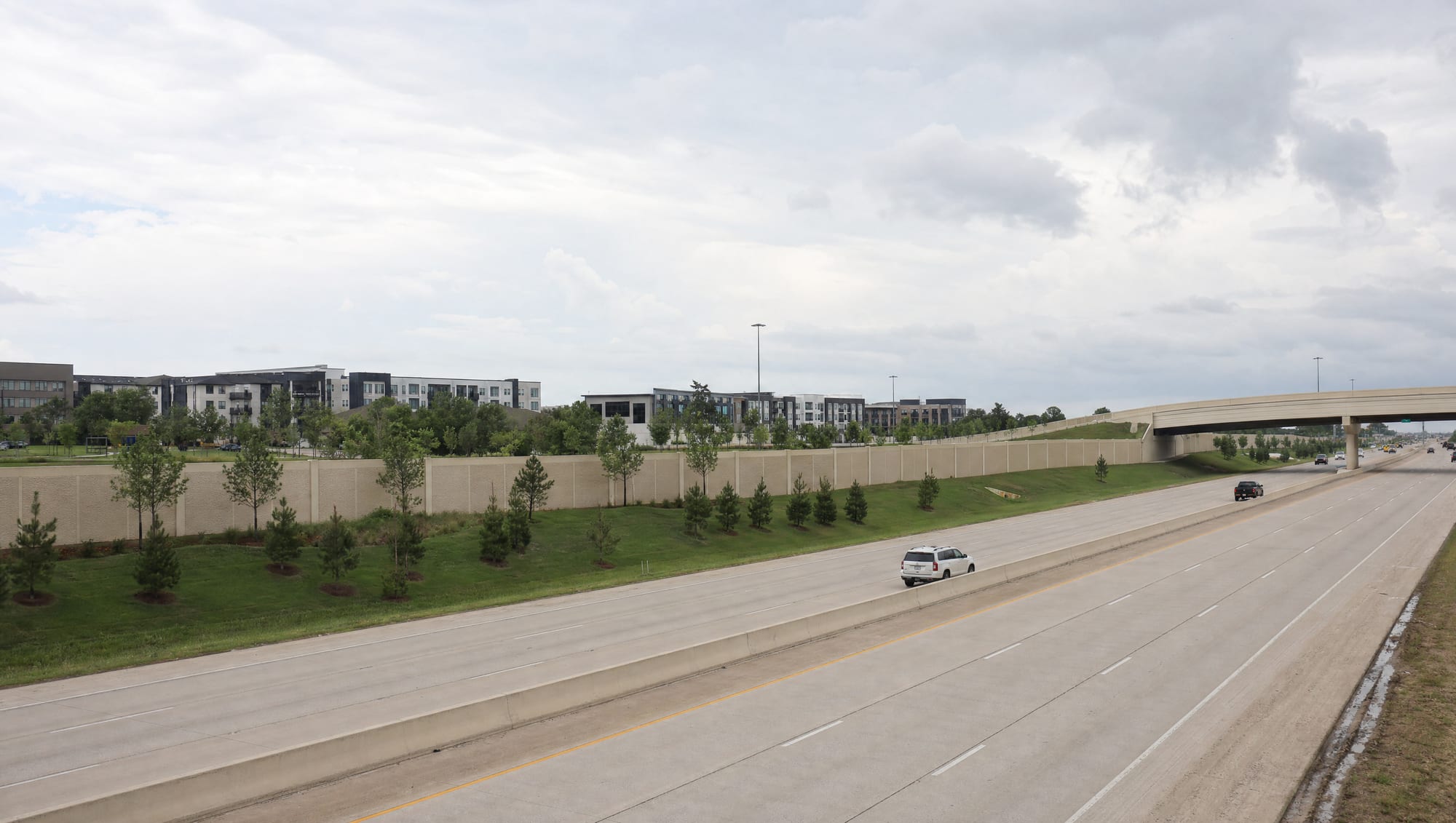 A wide multi-lane suburban highway corridor near Katy, Texas, with tall concrete sound walls, an overpass, landscaped grassy frontage, young trees, and master-planned West Houston development beyond under a big pale sky
