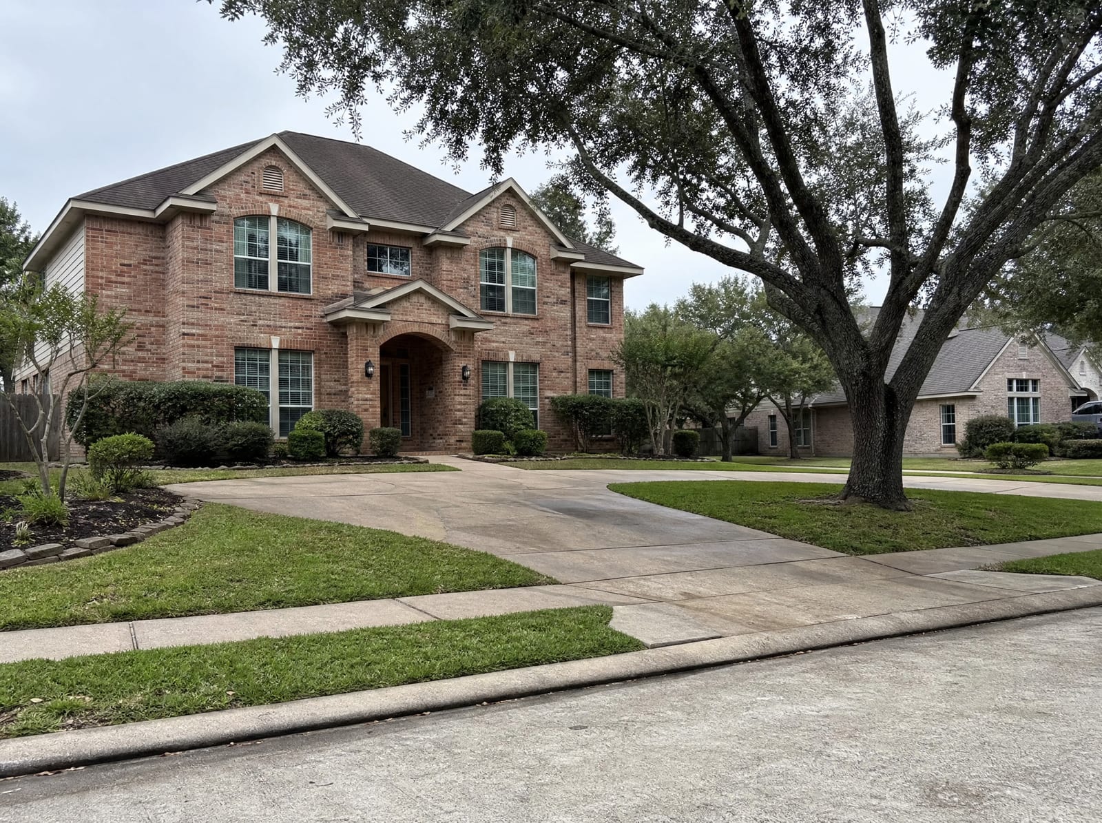 A quiet established residential street in Missouri City, Texas, with mature two-story brick suburban homes, grown shade trees, manicured lawns, wide sidewalks, and a curbed street under an overcast sky
