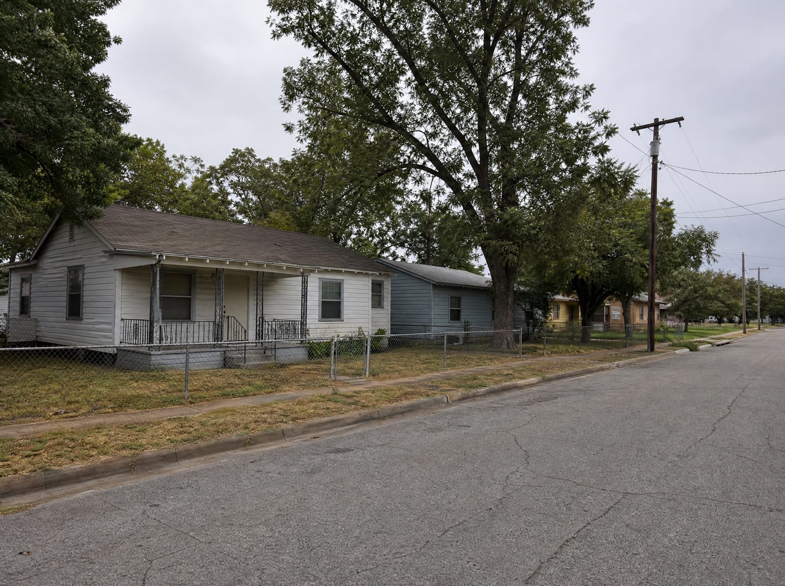 A modest working-class residential street in Pasadena, Texas, with older single-story homes, small front yards, mature shade trees, chain-link fences, and a cracked asphalt street under an overcast sky