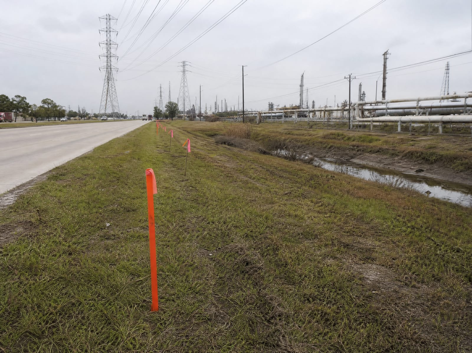 Orange survey stakes and pink right-of-way flags along a grassy strip beside a wide industrial arterial in Pasadena, Texas, with distant transmission towers, pipelines, and a drainage ditch, showing right-of-way acquisition pressure