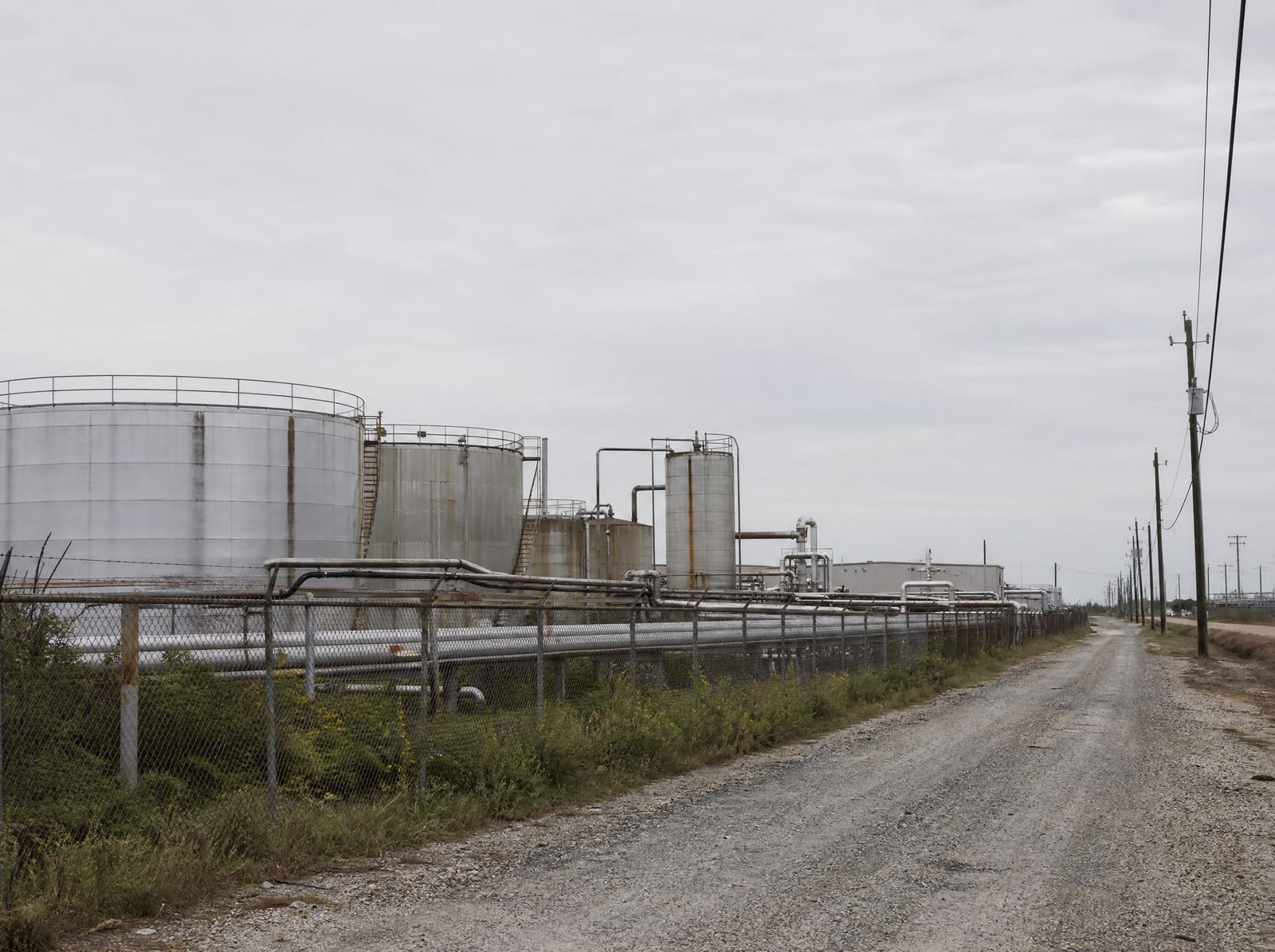 Industrial infrastructure near Pasadena, Texas, with silver storage tanks and a run of pipelines behind a chain-link fence, utility poles, and a gravel service road under a big pale overcast sky
