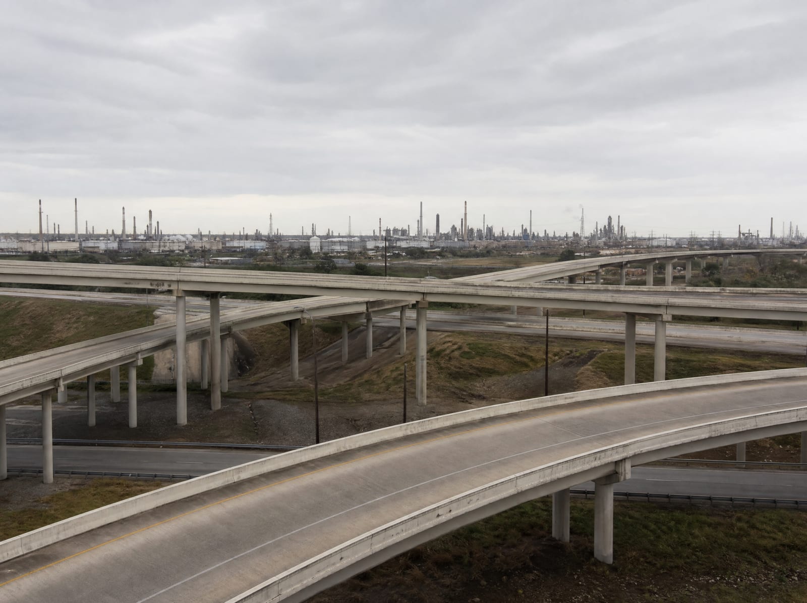 A wide highway interchange near Pasadena, Texas, with concrete overpasses and freeway ramps and an industrial refinery backdrop in the distance under a big pale overcast sky