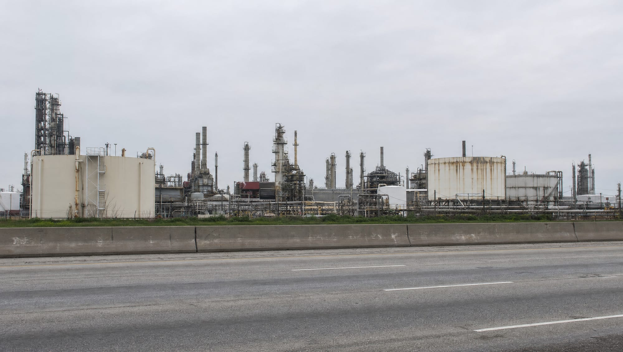 An industrial landscape near Pasadena, Texas along the Houston Ship Channel area, with petrochemical refinery towers, distillation columns, and large storage tanks beyond a multi-lane highway under a big pale overcast sky