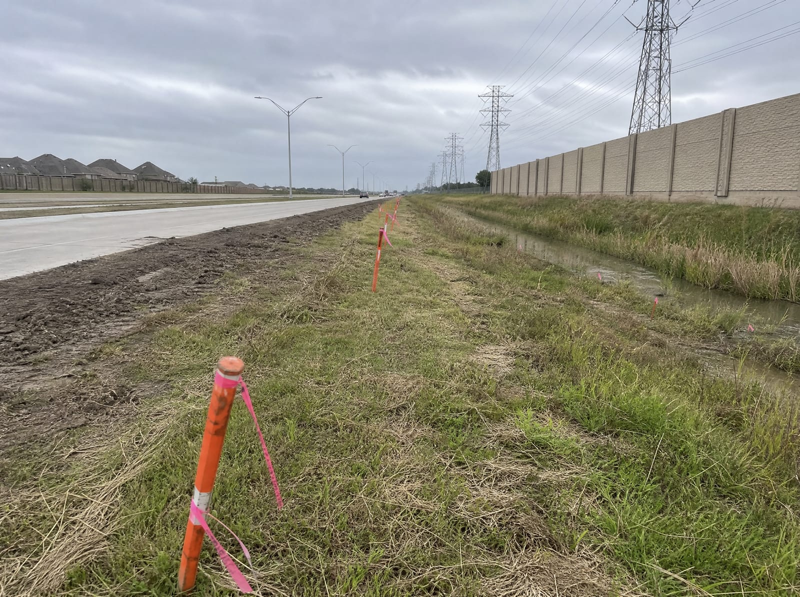 Orange survey stakes and pink right-of-way flags along a grassy strip beside a widening multi-lane suburban arterial in Pearland, Texas, with a concrete sound wall and distant transmission towers, showing right-of-way acquisition pressure