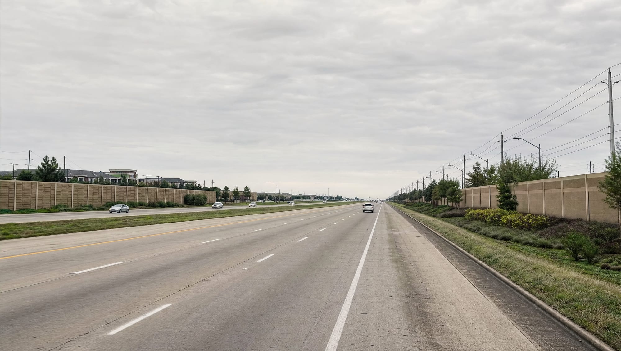 The State Highway 288 corridor running through Pearland, Texas, a wide divided multi-lane highway with sound walls, landscaped frontage, utility lines, and fast-growing suburban commercial development beyond under a big pale sky