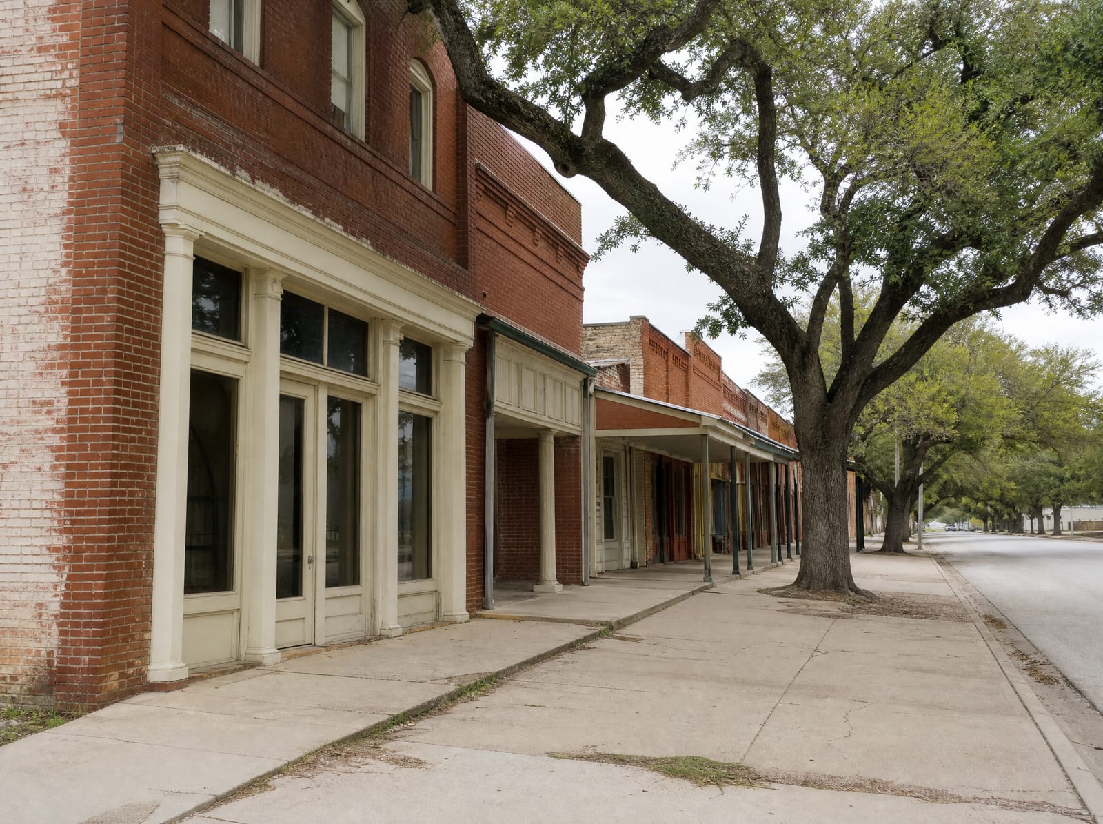 A quiet historic small-town Texas main street of modest brick storefronts lined with mature live oaks under a soft overcast sky, evoking the Richmond county seat