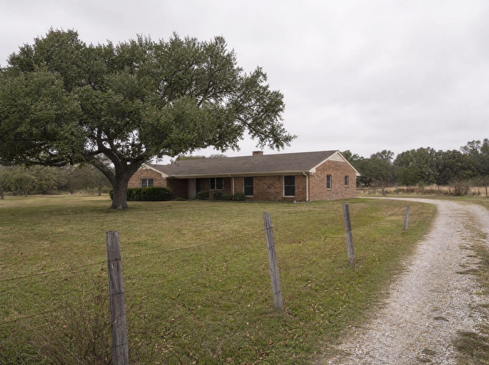 A modest single-story Texas brick ranch-style home on a grassy Fort Bend County lot with a live oak and a wire-and-post fence line under a big pale overcast sky near Richmond, Texas