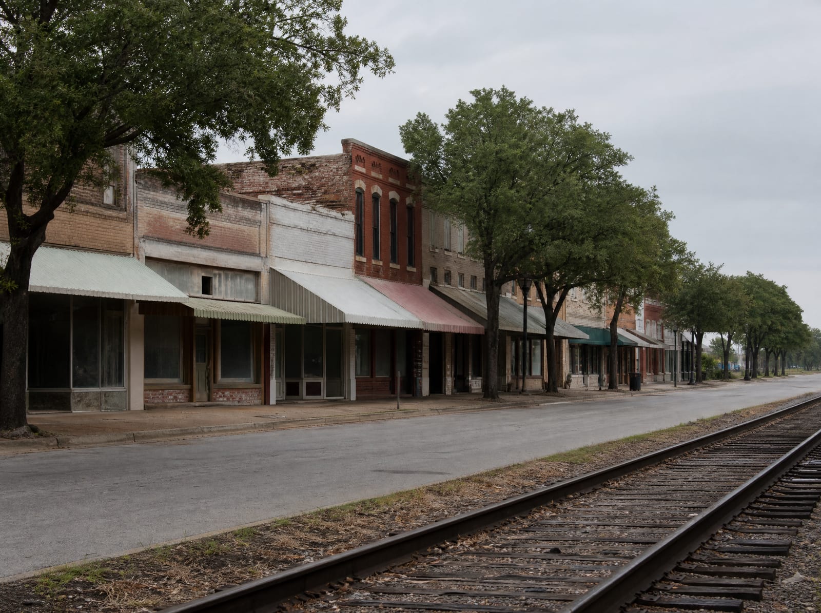 A quiet historic downtown main street in Rosenberg, Texas, with a row of weathered early-twentieth-century brick storefronts beside a railroad track, mature street trees, and wide sidewalks under an overcast sky