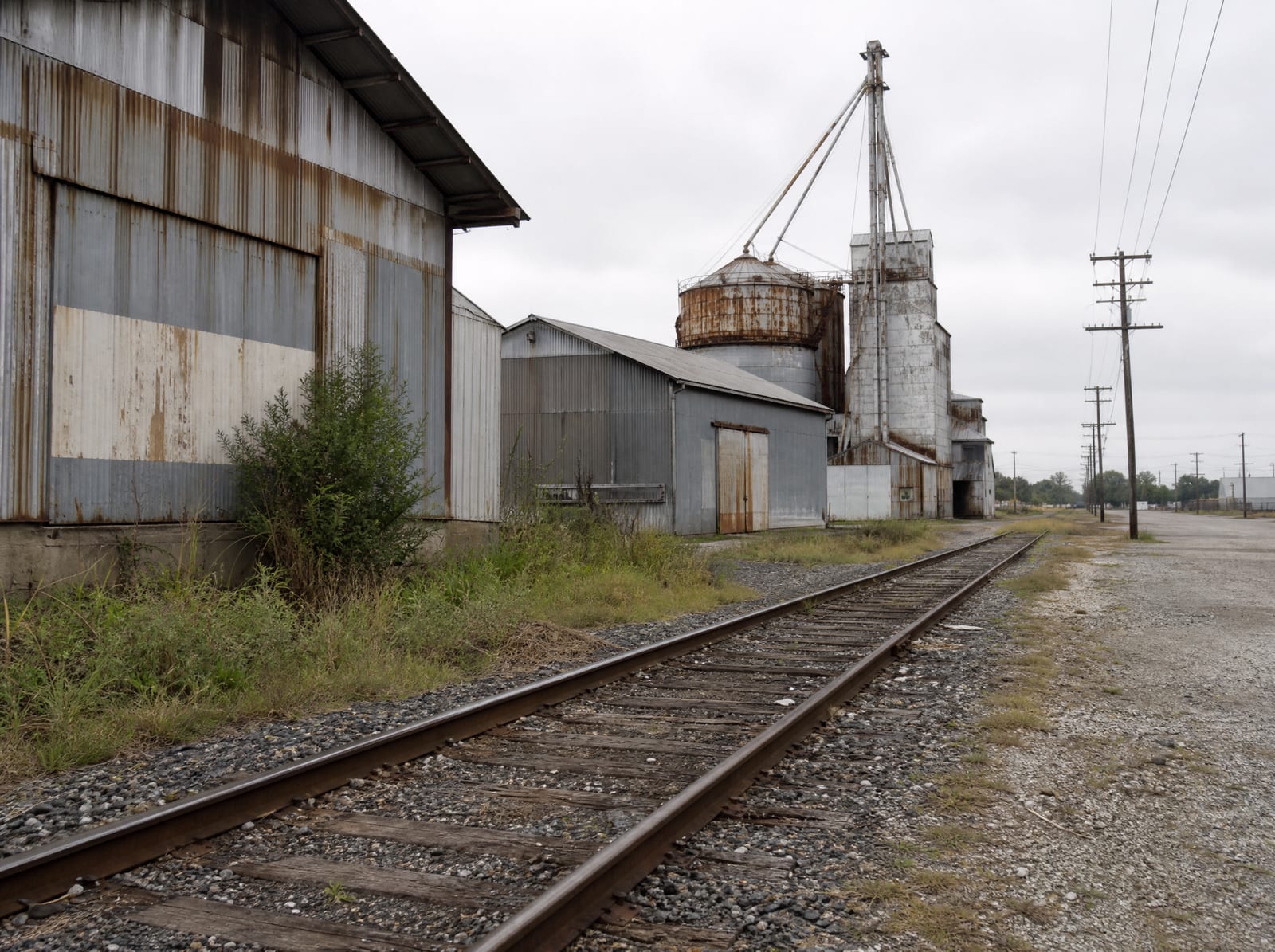 A single railroad track running past older weathered metal warehouses and a grain elevator in a rail-side industrial district of Rosenberg, Texas, with a gravel lot and utility poles on flat Fort Bend terrain