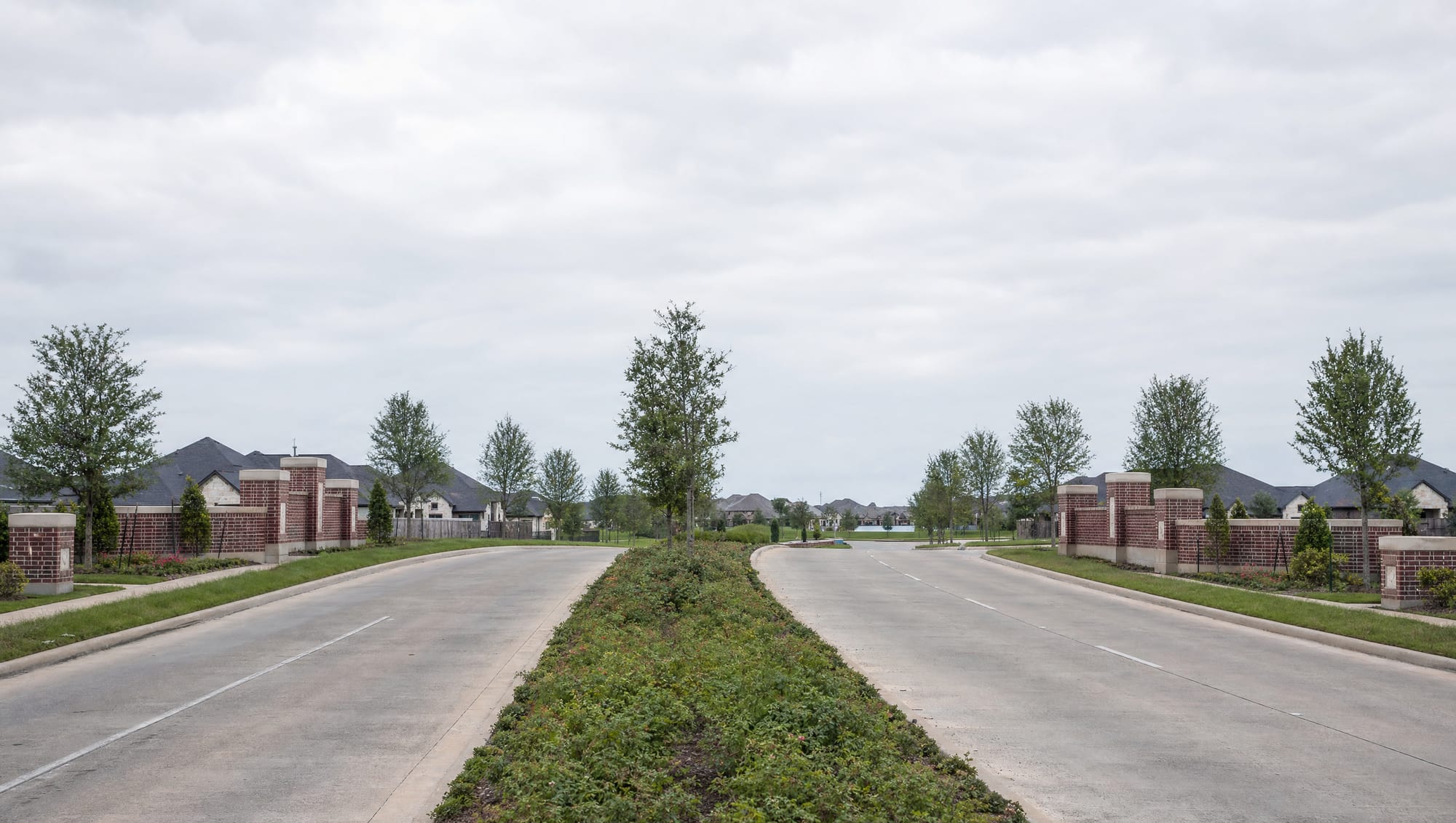 A landscaped master-planned community entrance boulevard in Sienna near Missouri City, Texas, a wide divided road with a planted median, young trees, low brick monument walls, and newer suburban development beyond under a big pale sky