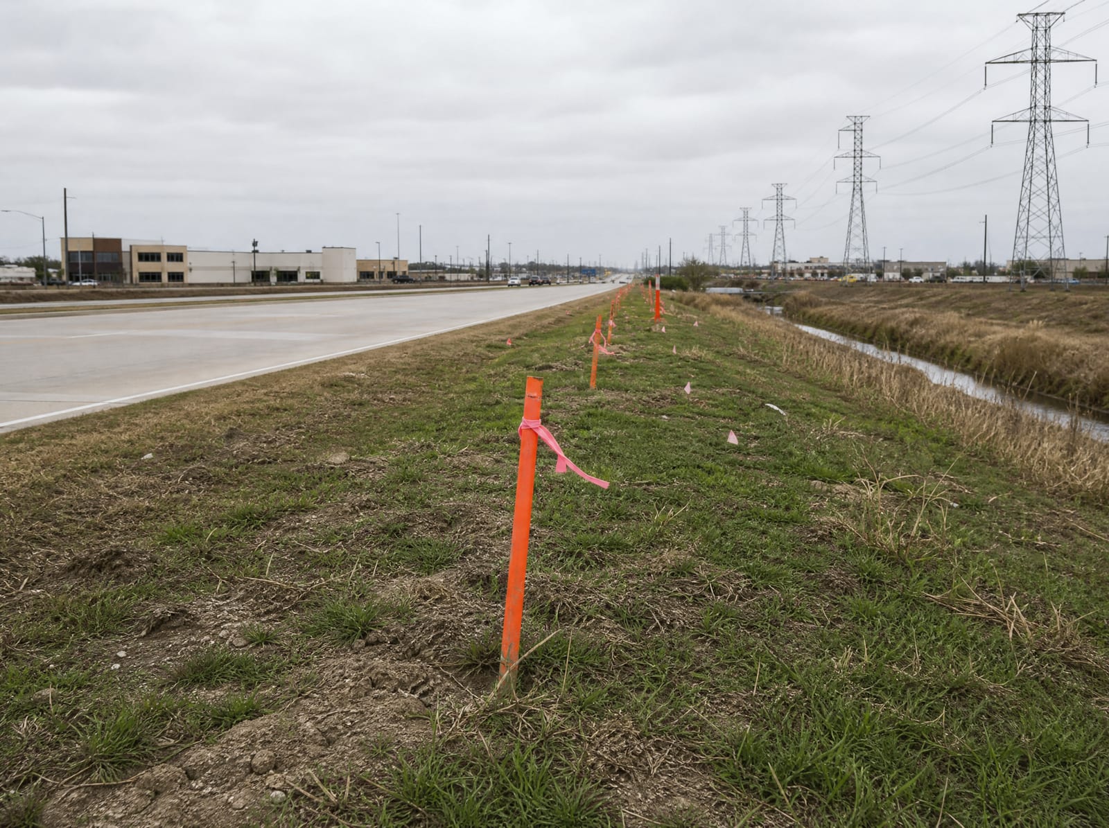 Orange survey stakes and pink right-of-way flags along a grassy strip beside a widening commercial arterial in Stafford, Texas, with distant transmission towers and a drainage ditch, showing right-of-way acquisition pressure