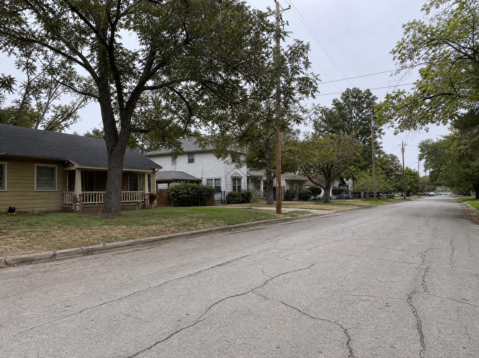 A quiet established modest residential street in Stafford, Texas, with older single-story and two-story homes, mature shade trees, front lawns, and a cracked asphalt street under an overcast sky