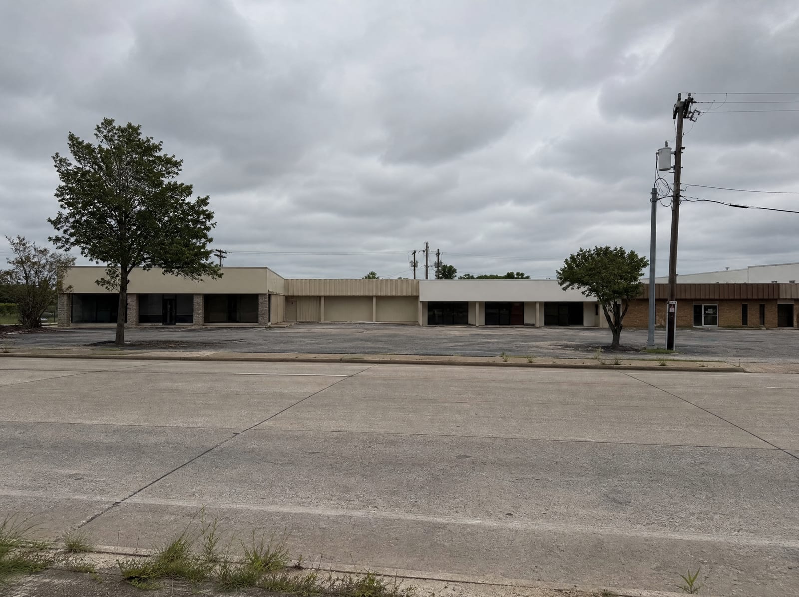 A modest commercial frontage strip in Stafford, Texas, with low one-story retail and office buildings, a shared asphalt parking lot, small trees, and utility poles along a wide arterial road under an overcast sky