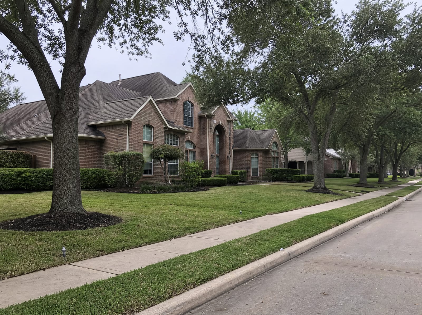 A quiet established affluent residential street in Sugar Land, Texas, with large two-story brick homes, mature manicured landscaping, tall shade trees, wide sidewalks, and a curbed street under an overcast sky
