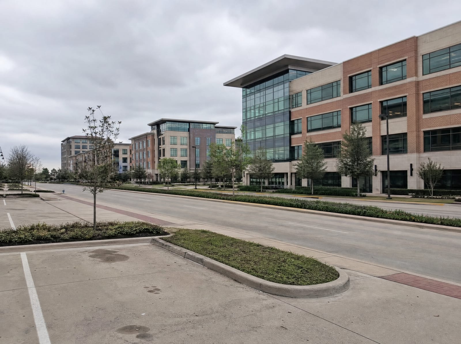 A modern low-rise office and commercial district in Sugar Land, Texas, with contemporary brick and glass mid-rise buildings, landscaped medians and parking, and young trees along a wide boulevard under an overcast sky