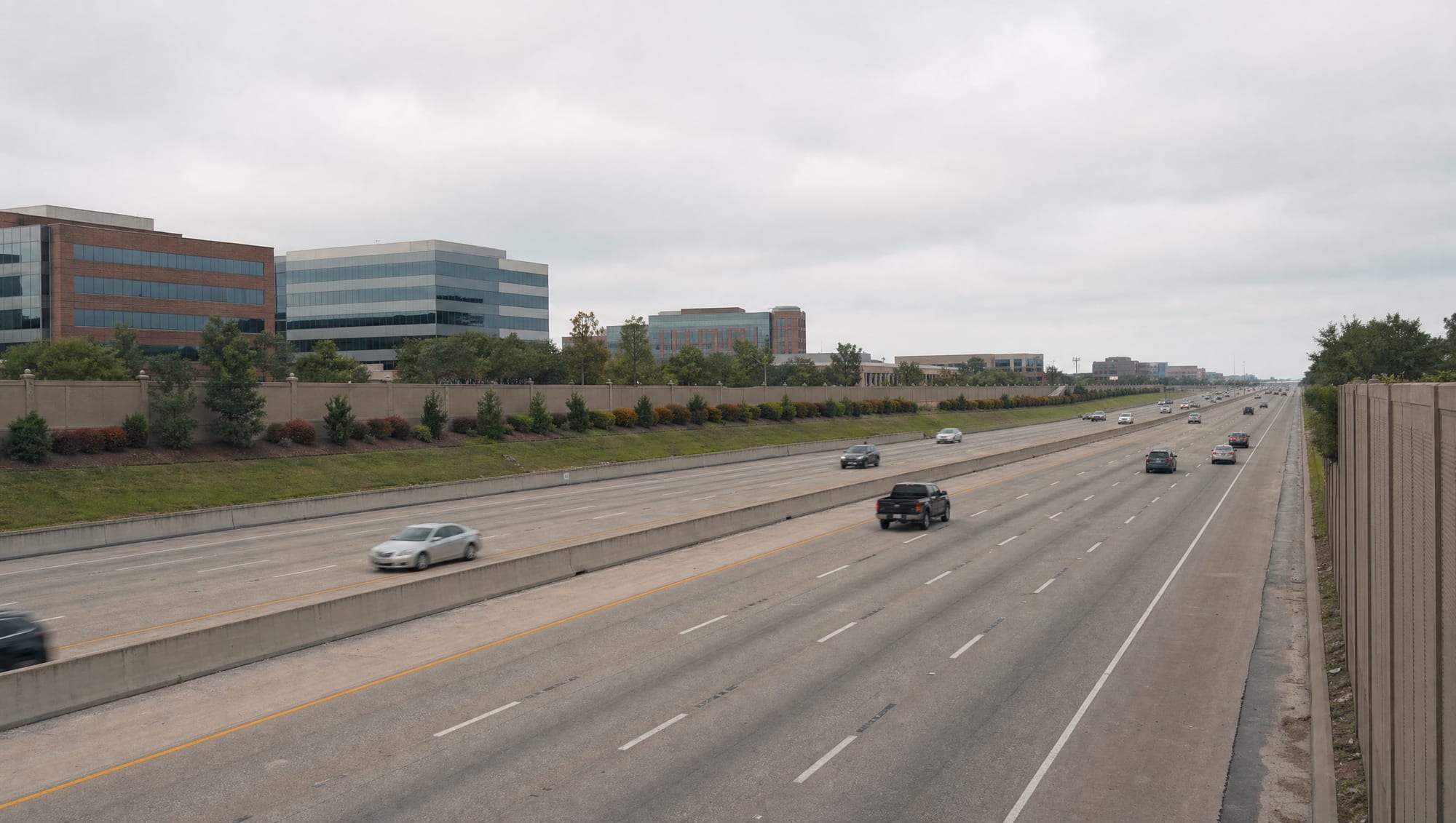 The US-59 Southwest Freeway corridor running through Sugar Land, Texas in Fort Bend County, a wide modern multi-lane highway with sound walls, landscaped frontage, and low-rise office and commercial buildings beyond under a big pale sky