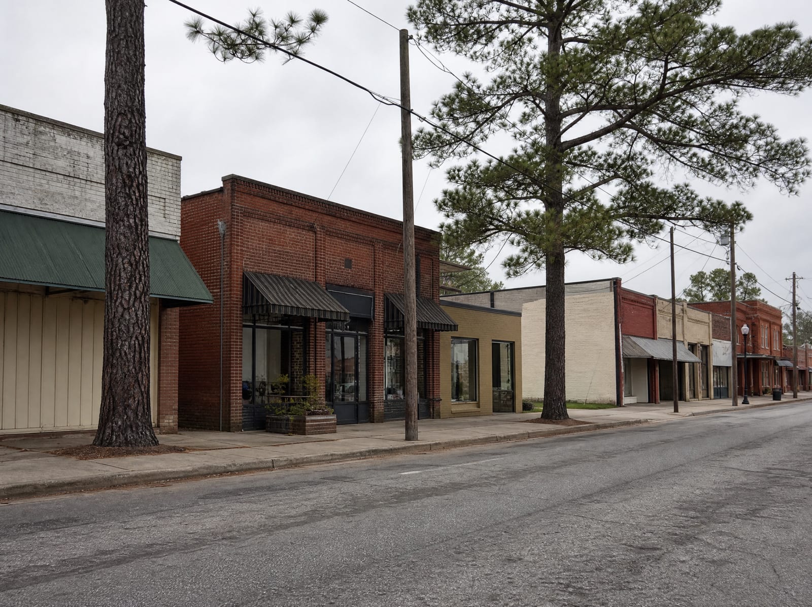 A quiet older small-town street in Tomball, Texas, with modest one and two-story brick storefronts and low commercial buildings among tall pine trees, wide sidewalks, and utility poles under an overcast sky